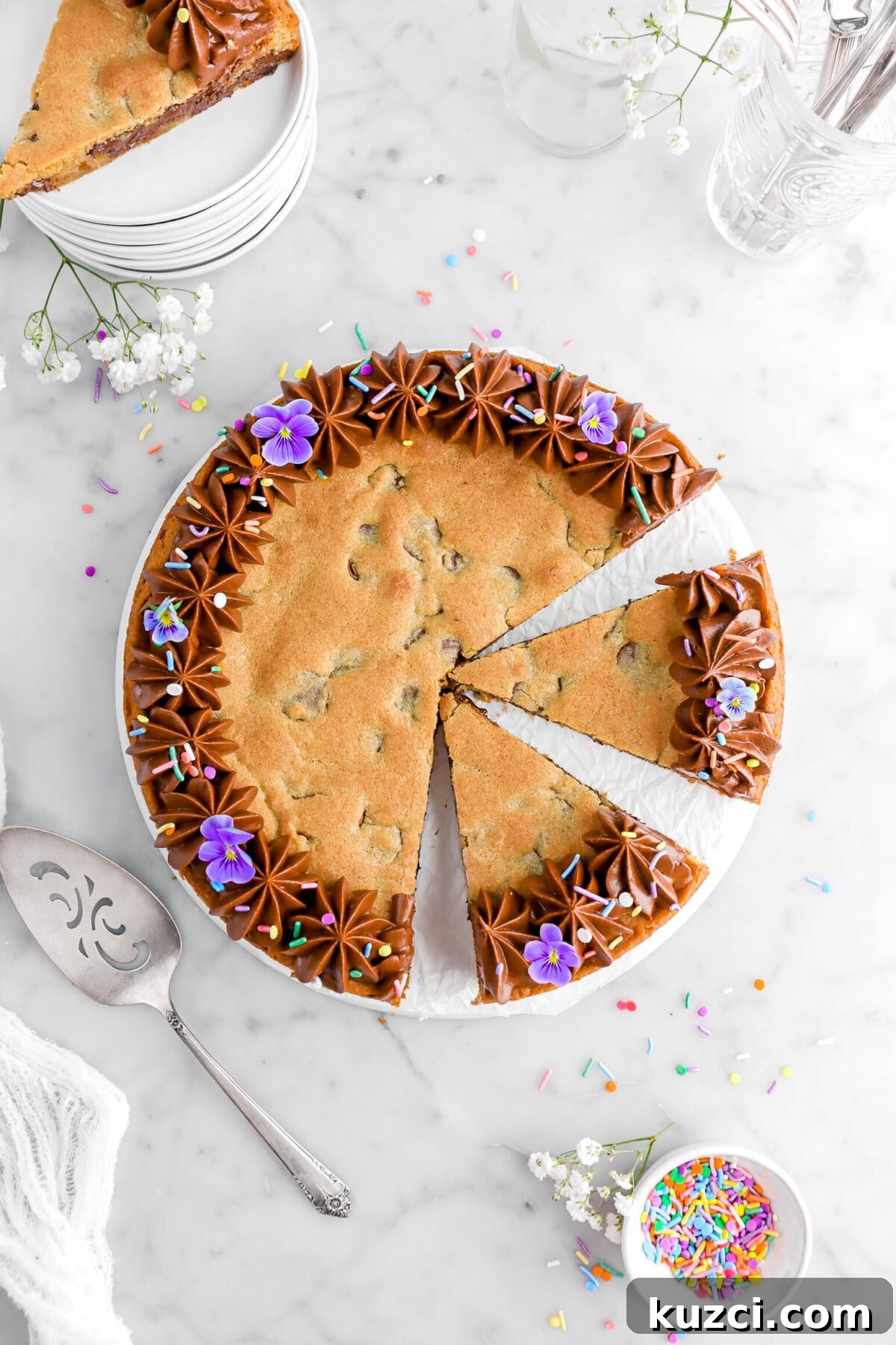 Chocolate Chip Cookie Cake with Chocolate Buttercream 5 overhead shot of chocolate chip cookie cake with two slices cut into the cake with another slice on stack of plates above, a cake knife beside, with a small bowl of sprinkles, and white flowers.