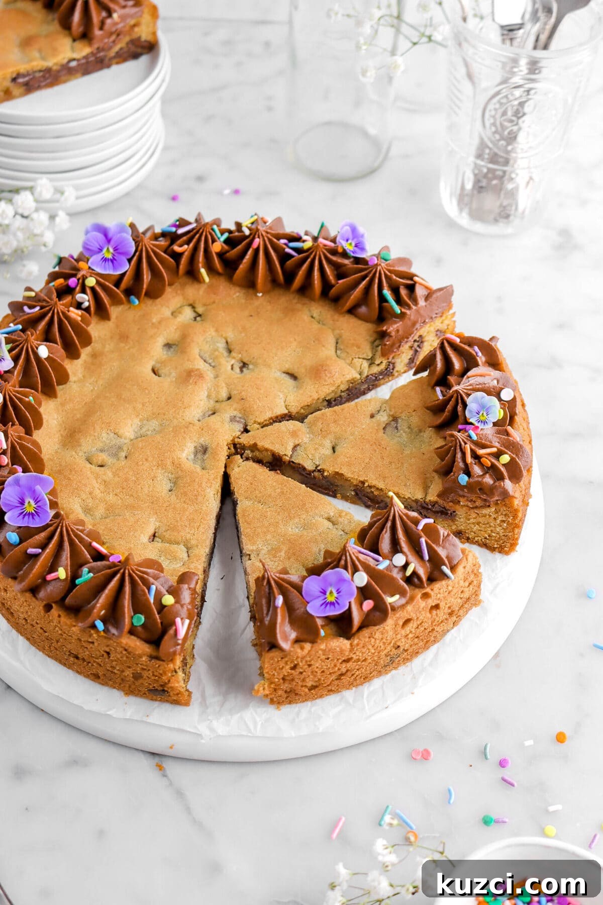 Chocolate Chip Cookie Cake with Chocolate Buttercream 6 cropped angled close up of chocolate chip cookie cake with two slices cut into it, chocolate frosting piped around the edges, with sprinkles, and purple flowers.