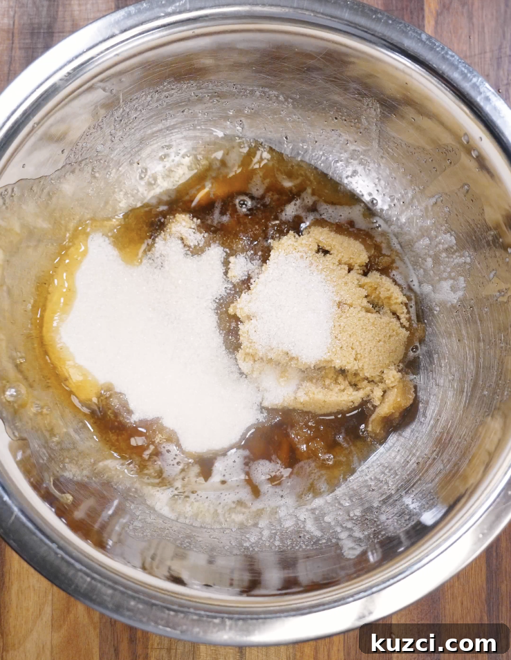 Warm brown butter combined with granulated sugar and dark brown sugar in a stainless steel mixing bowl before whisking to create the cookie dough base.