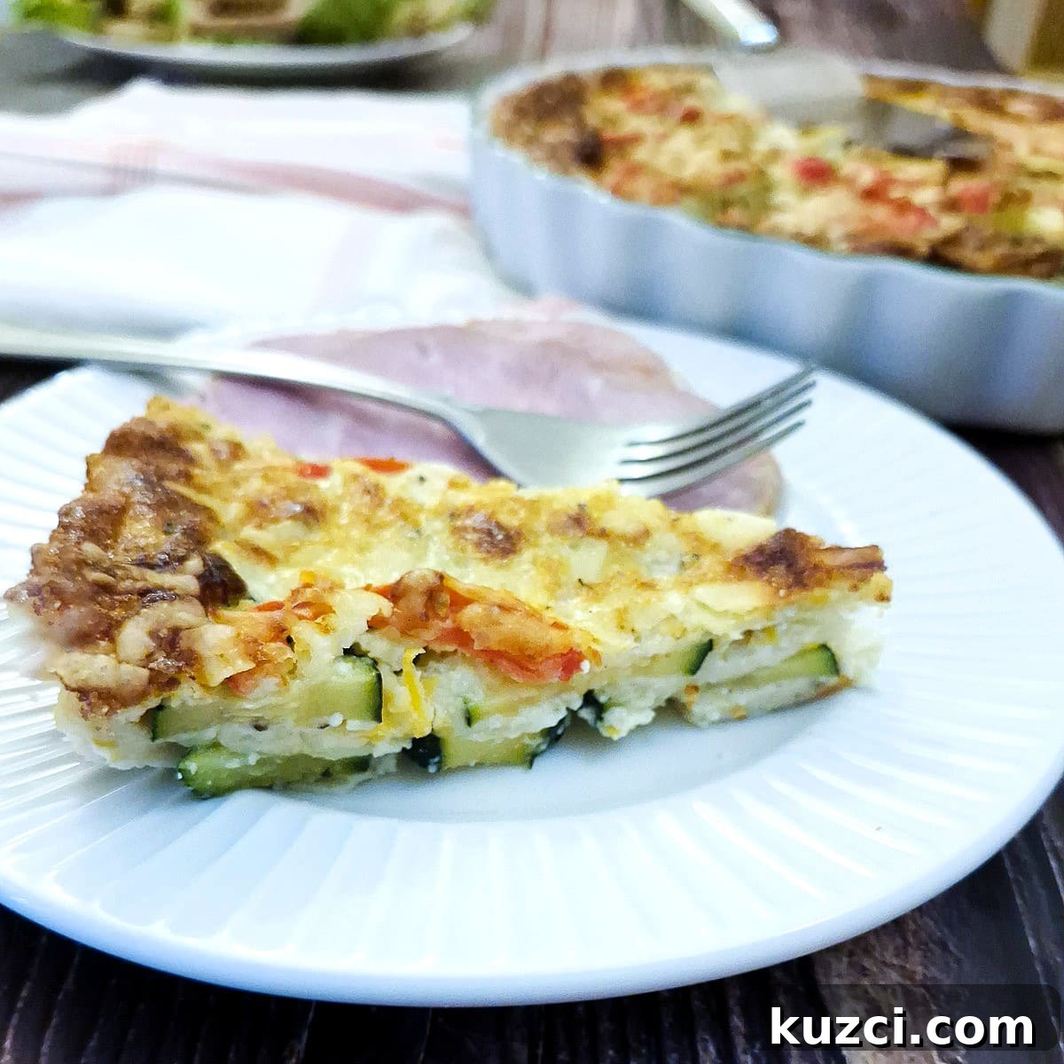 Crustless Cheesy Zucchini and Carrot Quiche 15 Closeup of a slice of courgette and carrot quiche on a white plate with a slice of gammon and a fork.