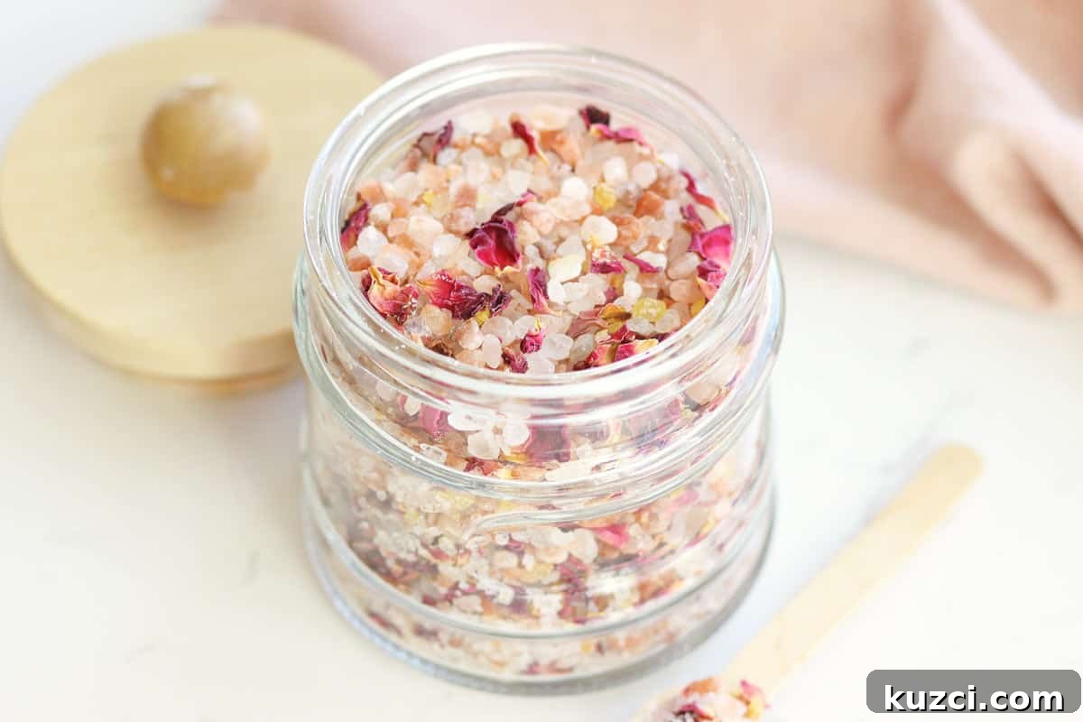 Homemade Rose Petal Bath Salts Recipe 4 a small glass jar with a wooden lid and small wooden spoon full of rose bath salts with rose petals in it.