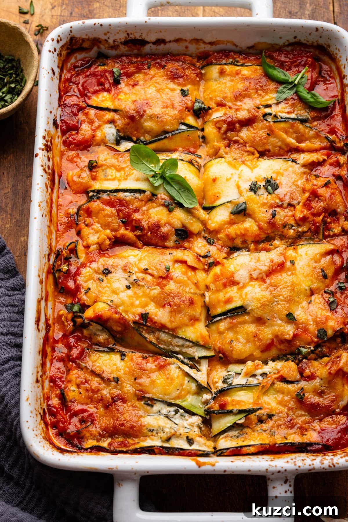 Zucchini Lasagna Recipe with Ricotta and Fresh Basil 7 A rectangle-shaped white baking dish with zucchini lasagna in it sliced into eight pieces. The dish is on a wooden board with fresh herbs and a dark gray napkin decorated around it.