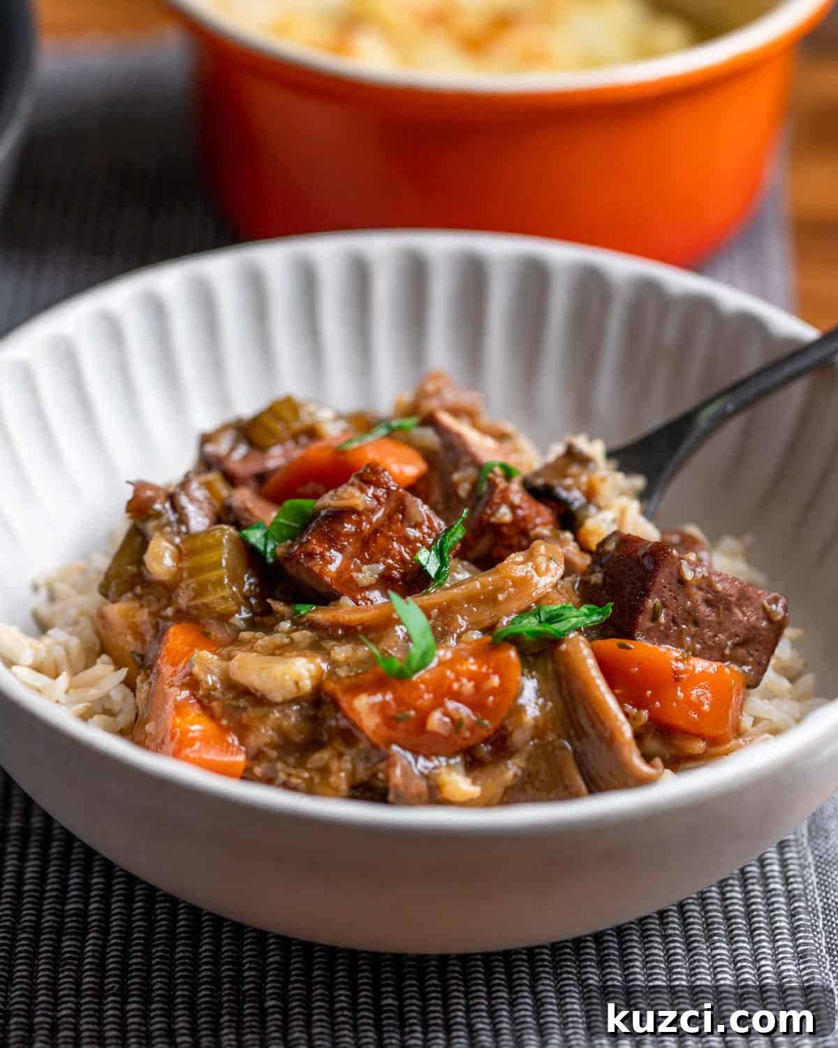 Vegan Stew with Smoked Tofu and Vegetables 3 Photo of a bowl of vegan stew served with brown rice and a spoon, set on a grey mat with an orange pot in the background.