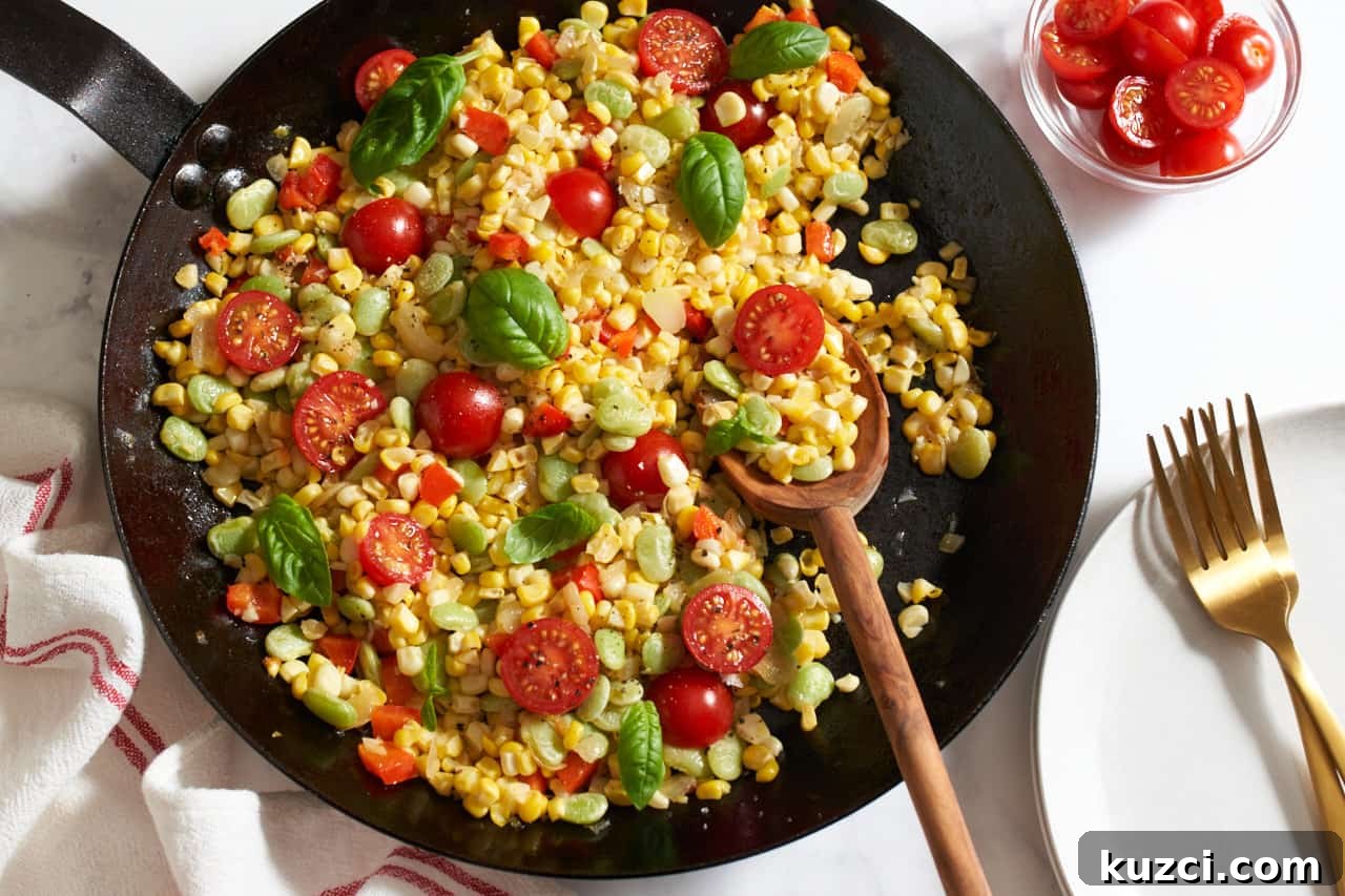 Sweet corn succotash topped with fresh basil in a black skillet with a wooden spoon in it. A red and white towel is in the bottom left, a plate with two gold forks is on the right, and a small bowl of sliced cherry tomatoes is in the top right corner.