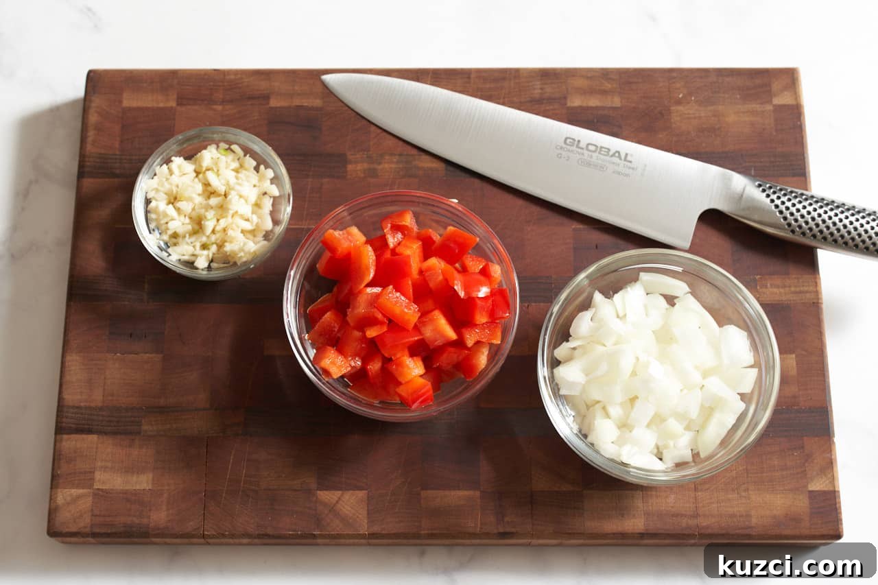 A knife on a wooden cutting board with three small glass bowls of diced garlic, red bell pepper, and onion.