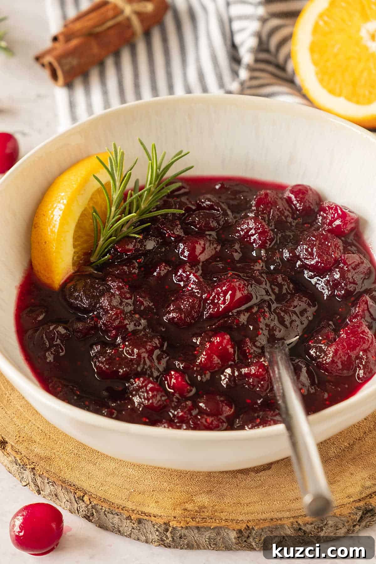 Homemade Cranberry Orange Sauce with Fresh Orange Juice 2 White bowl with cranberry sauce on a wooden round, orange slices, striped cloth in the background.