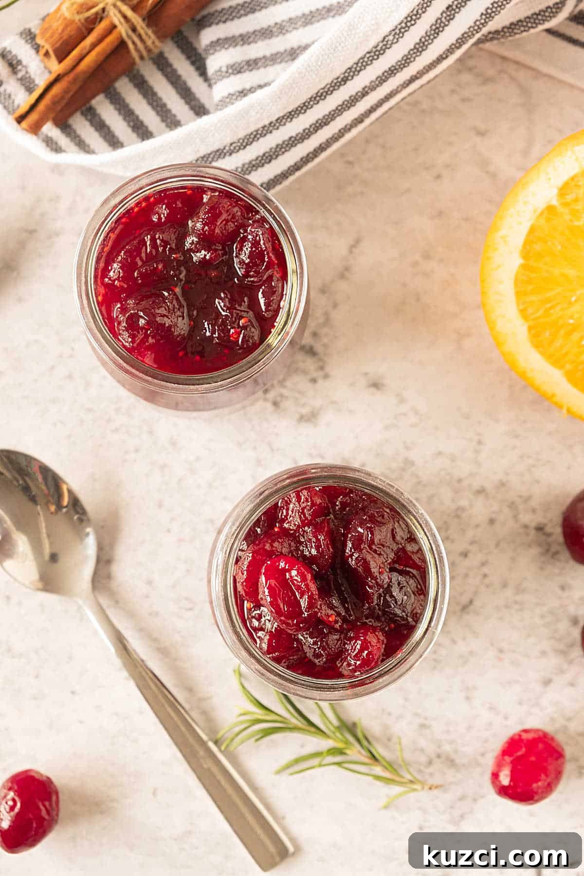 Homemade Cranberry Orange Sauce with Fresh Orange Juice 7 Top view of glass jars with cranberry sauce. Beige surface, striped cloth, orange pieces, silver spoon.