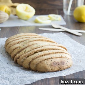 A loaf of bread with diagonal slashes sits on crumpled parchment paper atop a wooden table, with lemon halves, utensils, and a glass jar of water in the background.