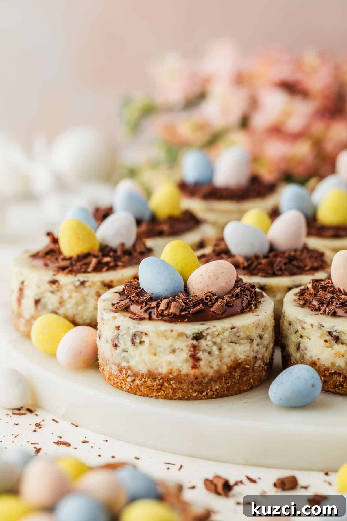 Mini Easter Egg Cheesecakes with Chocolate Ganache Recipe 2 A white platter of mini Easter egg cheesecakes next to a bowl of mini egg candies and pink flowers on a white backdrop.