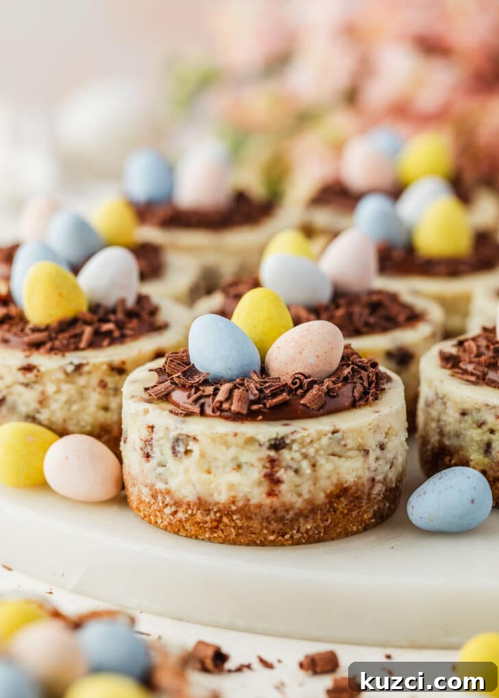 Mini Easter Egg Cheesecakes with Chocolate Ganache Recipe 18 A white platter of mini Easter egg cheesecakes on a tan counter next to pink flowers, a brown bowl of candies, and white eggs.