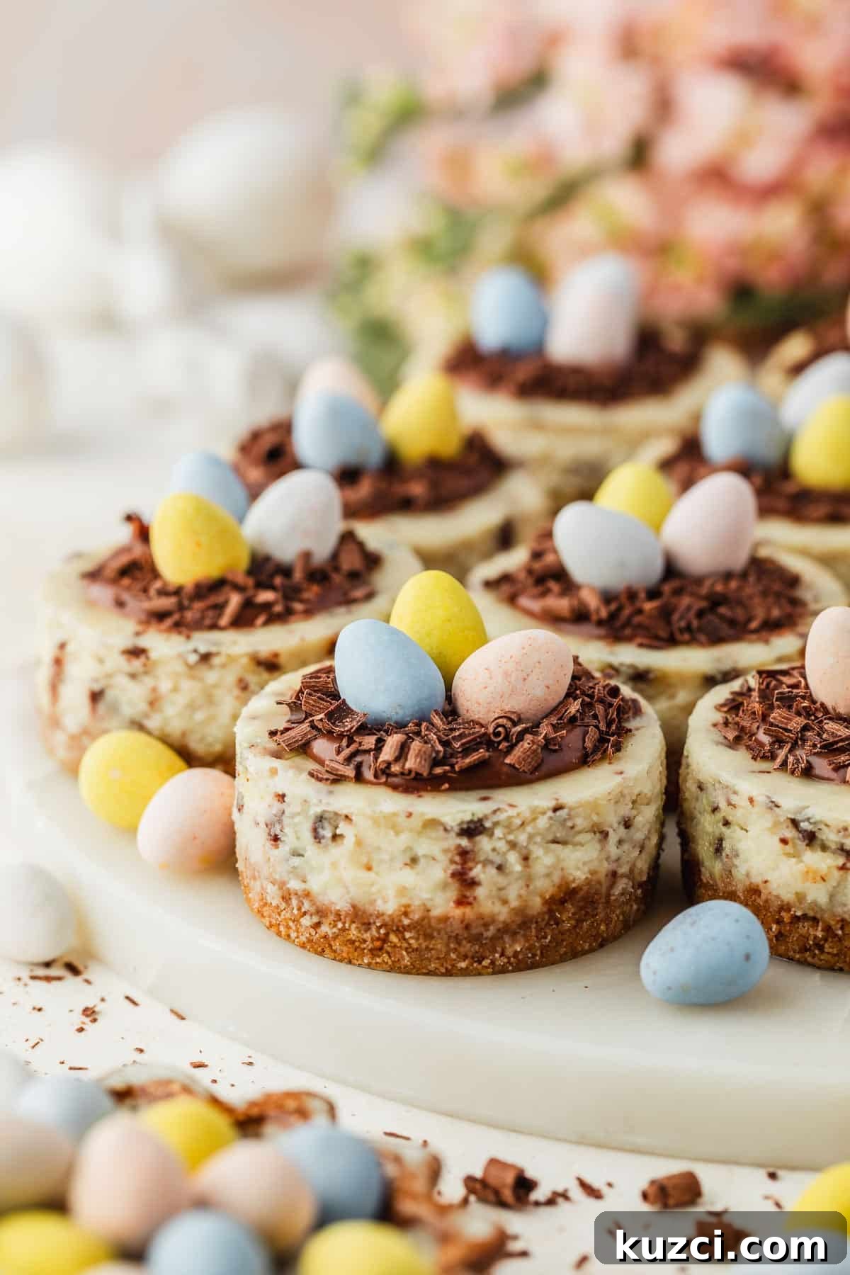 Mini Easter Egg Cheesecakes with Chocolate Ganache Recipe 19 A marble platter of mini egg cheesecakes with ganache nests next to a brown bowl of candies, pink flowers, and white ceramic bunnies on a white counter.