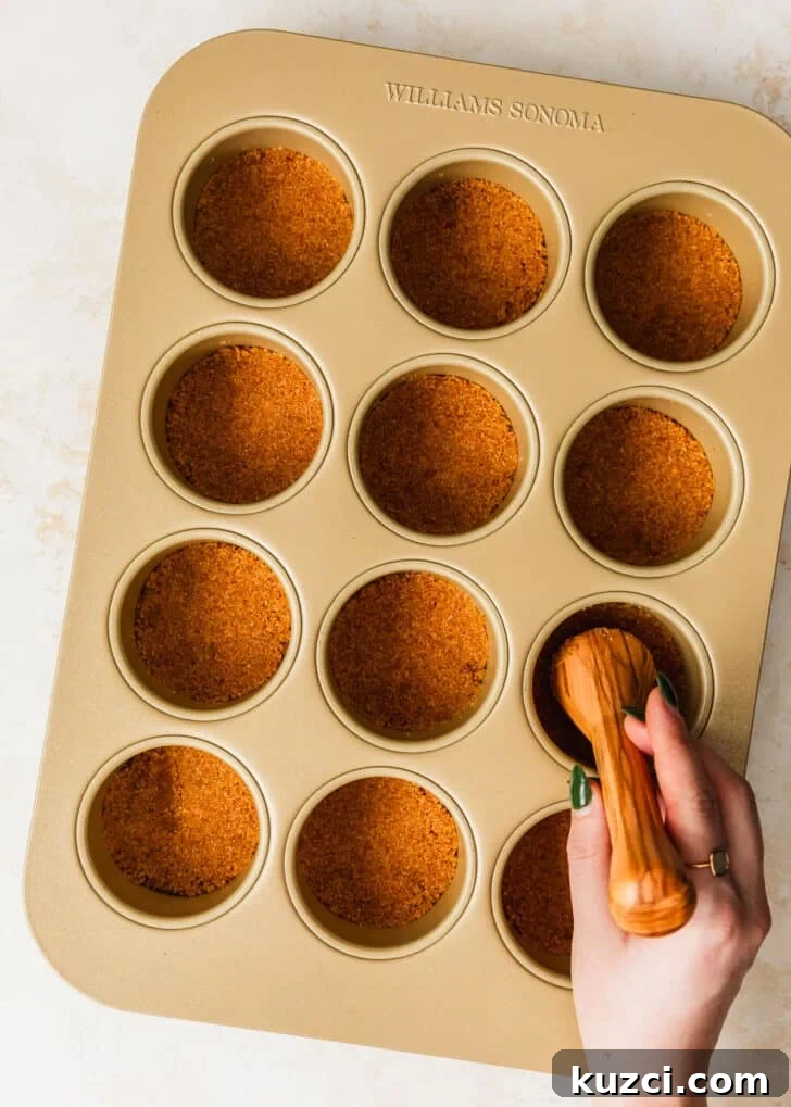 Mini Easter Egg Cheesecakes with Chocolate Ganache Recipe 5 A hand using a pastry tamper to press graham cracker crust into a cupcake pan.