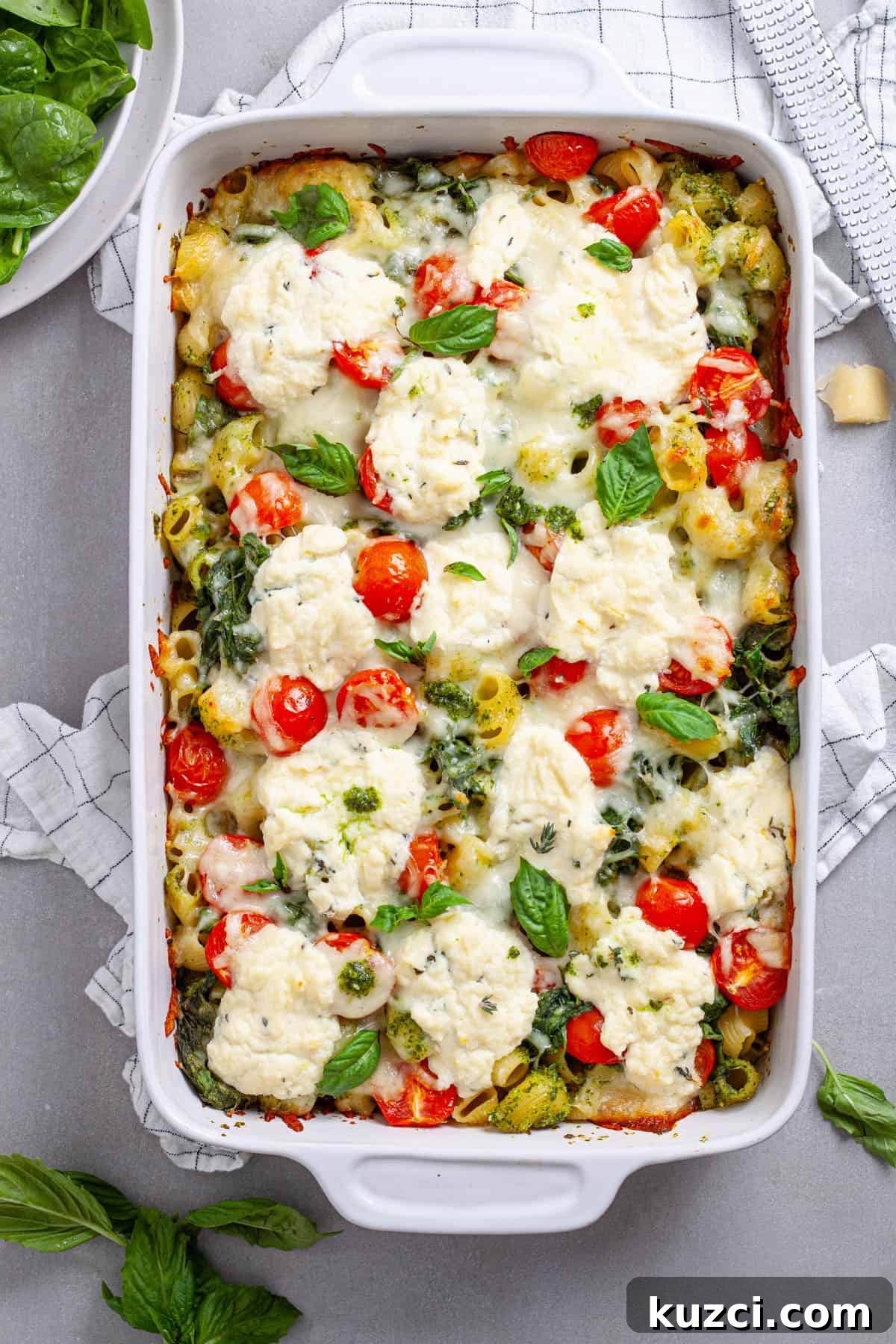 A casserole dish of ricotta pesto pasta with tomatoes and spinach on a gray table.