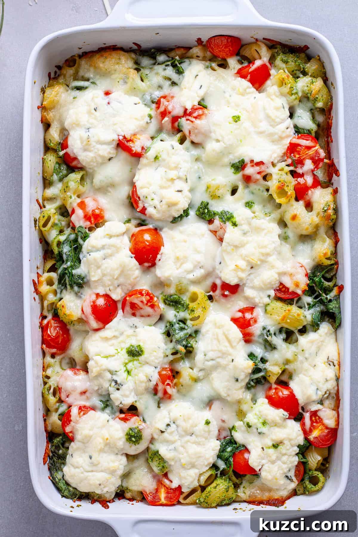 A casserole dish of pesto and ricotta baked pasta hot out of the oven topped with tomatoes and spinach.