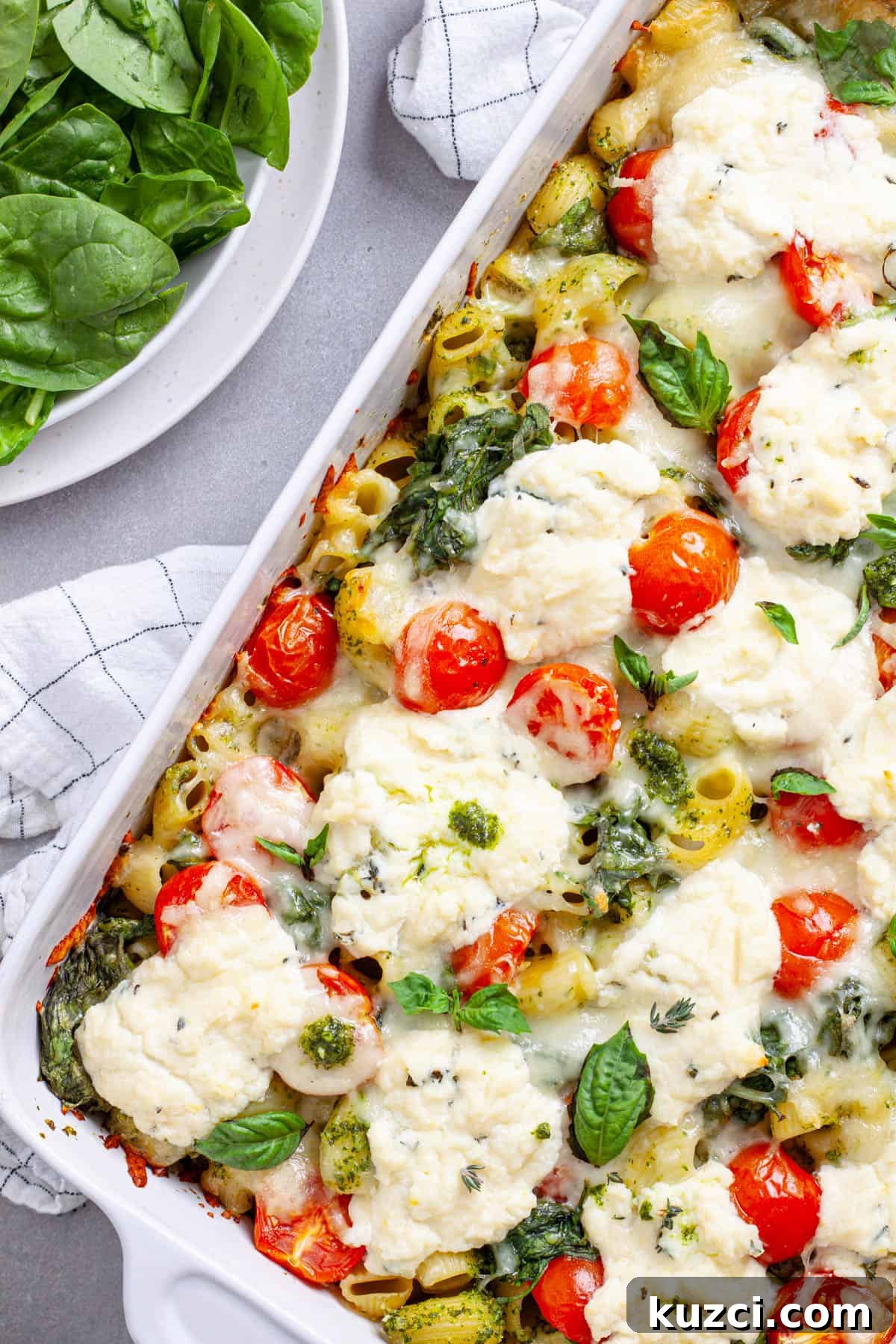A white casserole dish with ricotta pesto pasta topped with mozzarella, tomatoes and spinach.