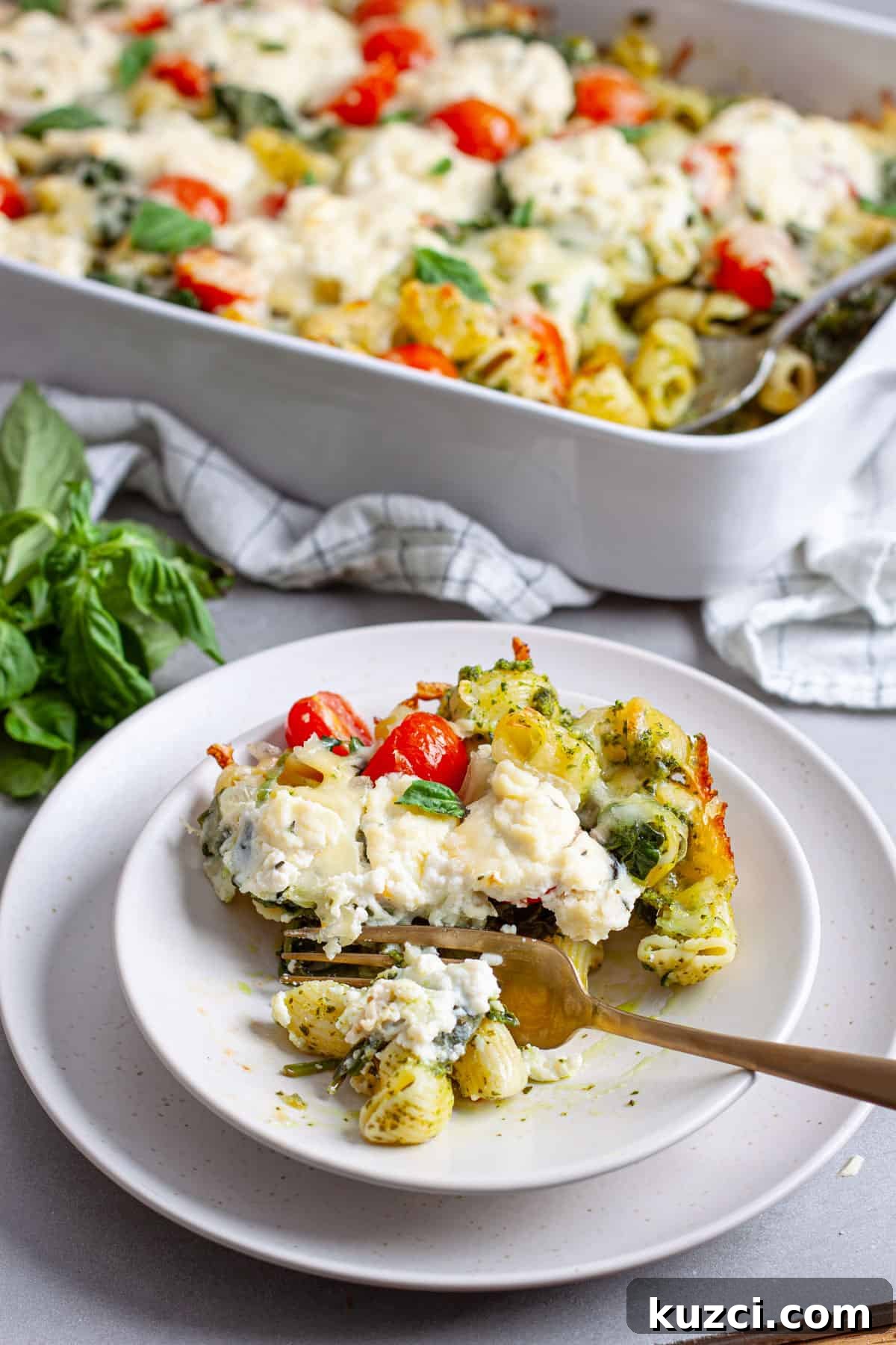 A serving of ricotta pesto pasta on a plate with a casserole dish of the baked pasta in the background.
