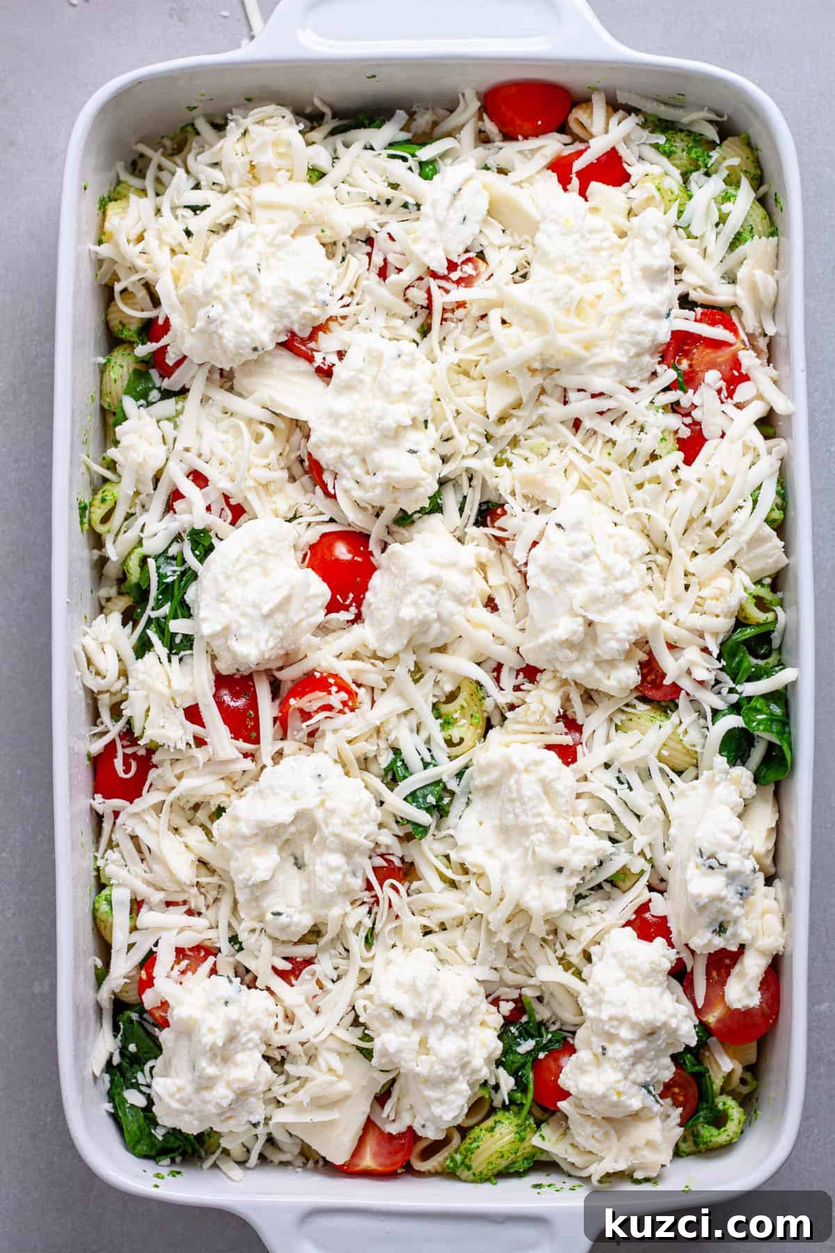 A casserole dish of pasta and pesto with ricotta and mozzarella scattered on top, along with tomatoes and spinach.