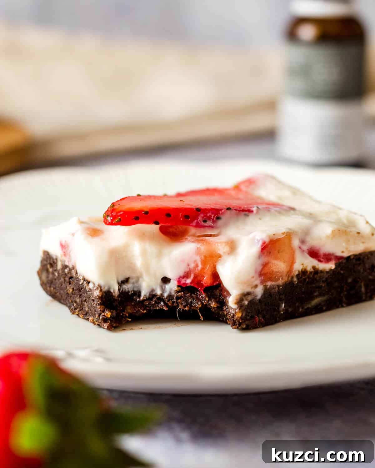 No-Bake Cheesecake Brownies Recipe 2 A cheesecake brownie with strawberries on a white plate. A bite has been taken. There is a bottle of CBD oil in the background.