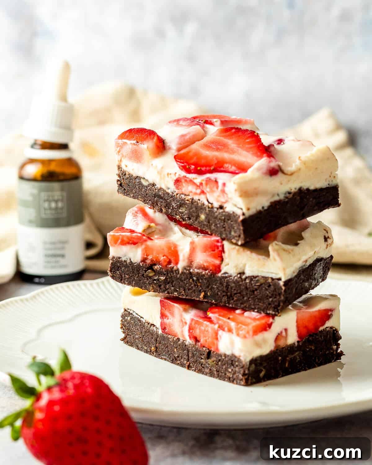No-Bake Cheesecake Brownies Recipe 3 Three cheesecake brownies with strawberries stacked on top of each other on a white plate. There is a strawberry as a prop and a bottle of CBD oil in the background.