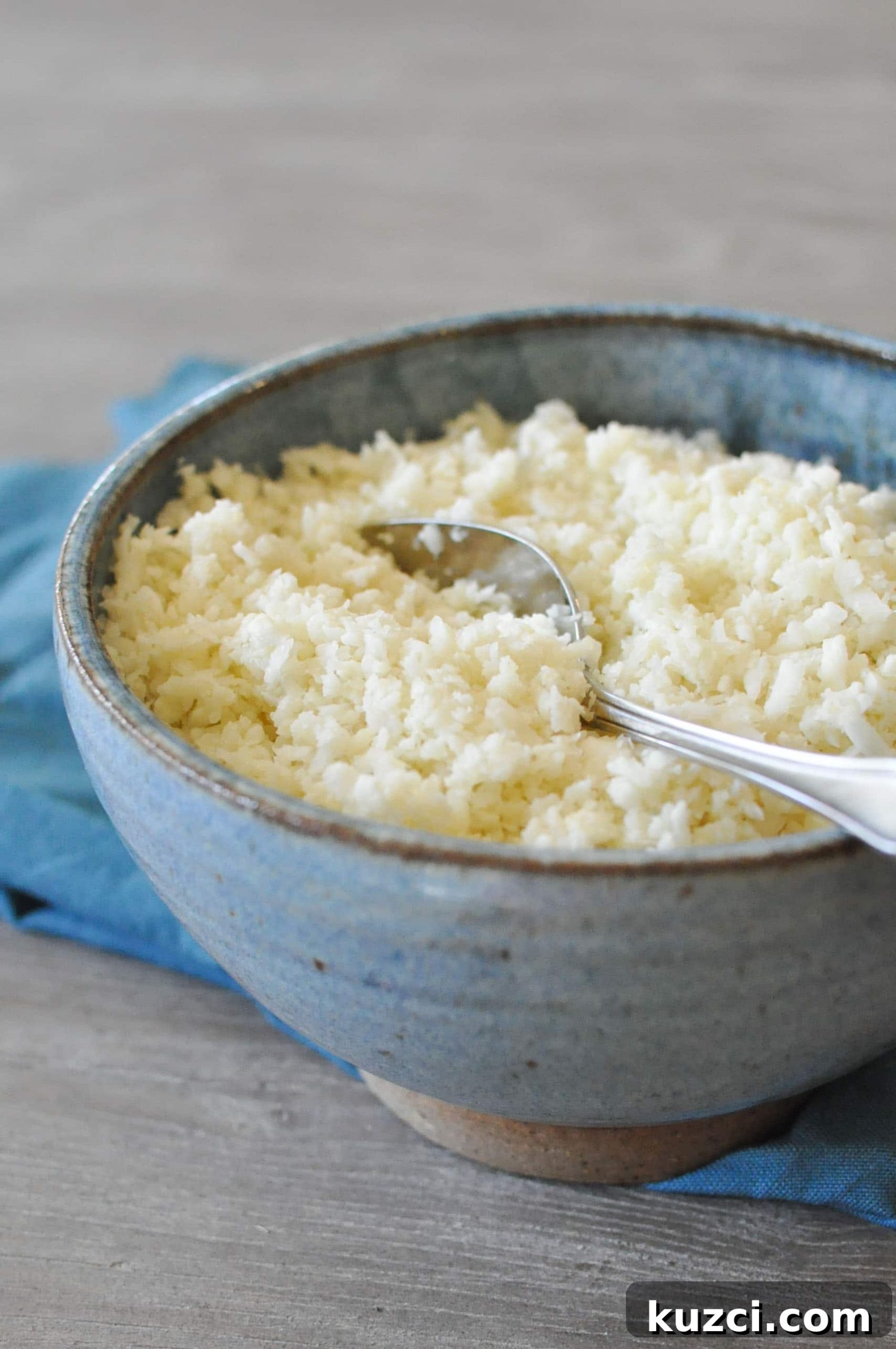 How to Make Cauliflower Rice 2 basic cauliflower rice in a blue bowl with a blue napkin on top of a grey wooden table