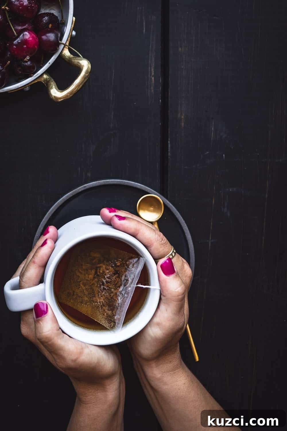 Hands with pink nails cradling a white mug of tea with a gold spoon on a dark background.