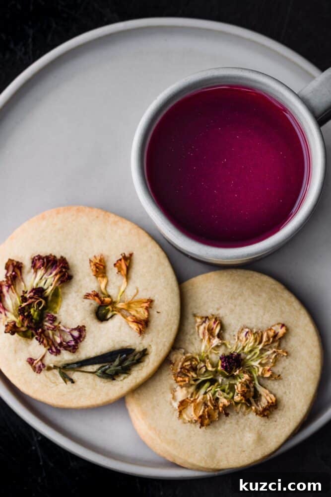 A burgundy tea in a white mug next to two dried edible flower shortbread cookies all on a light plate on a background,