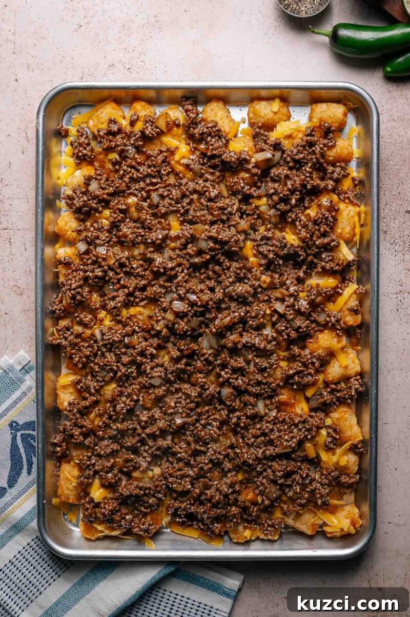A baking sheet topped with tater tots, melted cheese, ground beef, and onions, arranged on a kitchen towel next to whole jalapeños and a pepper shaker.