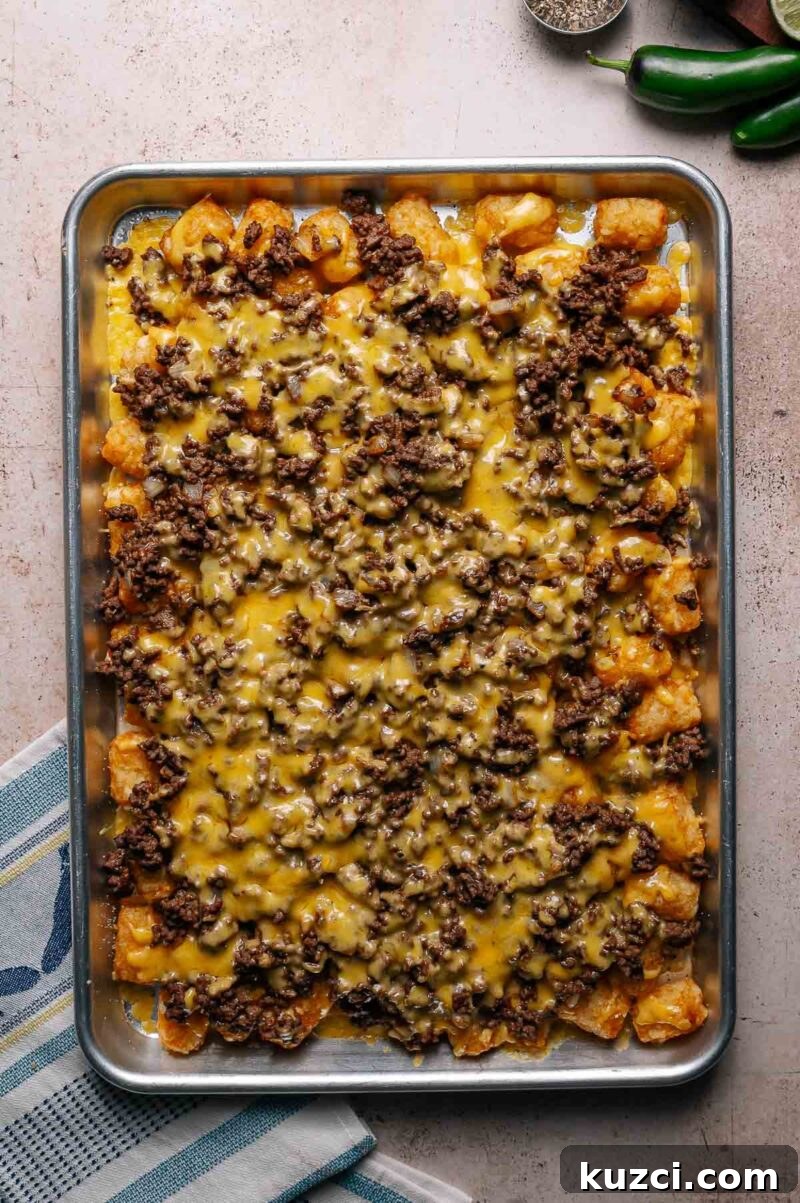 Rectangular baking tray filled with tater tots topped with cooked ground beef and melted cheese, photographed from above on a light-colored surface.