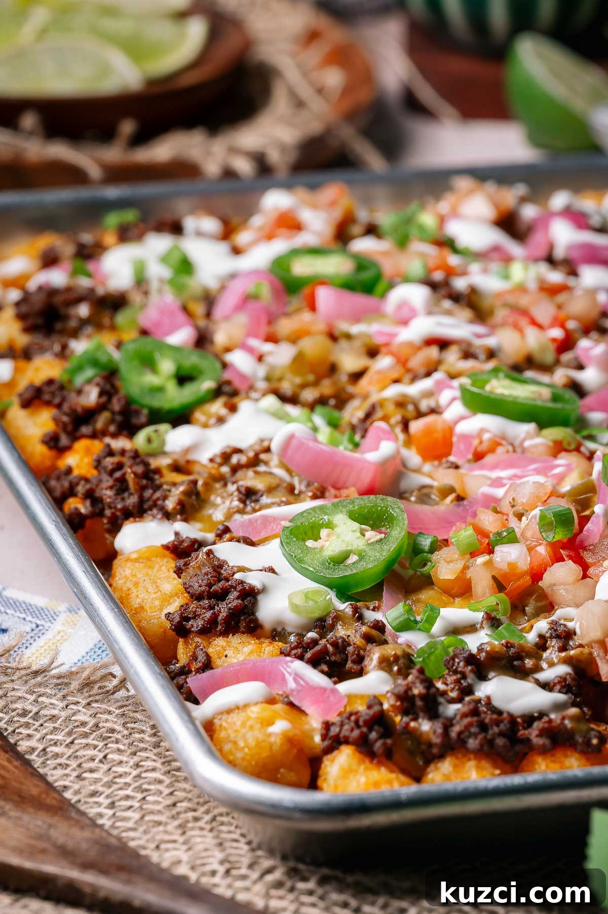 Sheet pan loaded tater tots topped with ground beef, melted cheese, jalapeño slices, diced tomatoes, green onions, pickled onions, and drizzled with sour cream.