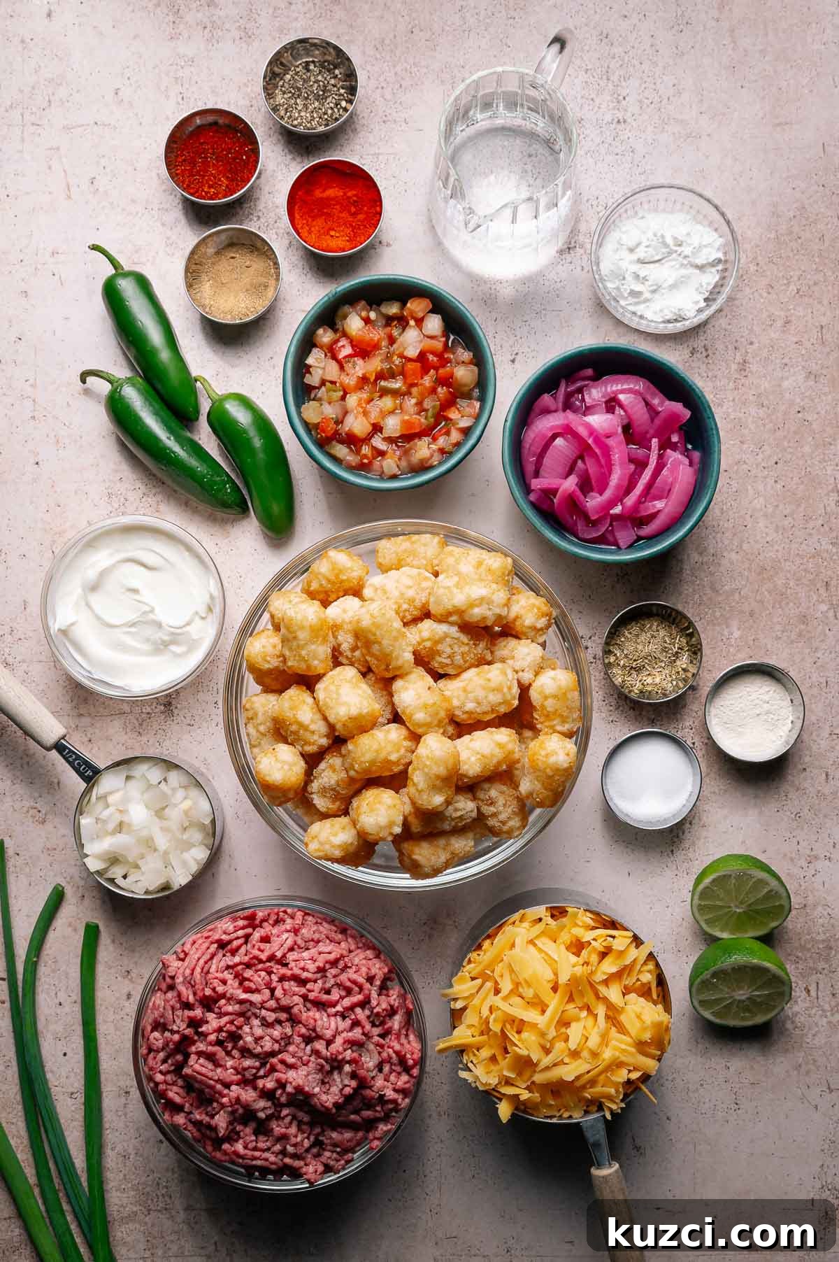 Ingredients for a loaded totchos laid out on a counter, including ground beef, cheese, tater tots, jalapeños, spices, sour cream, limes, and various toppings.