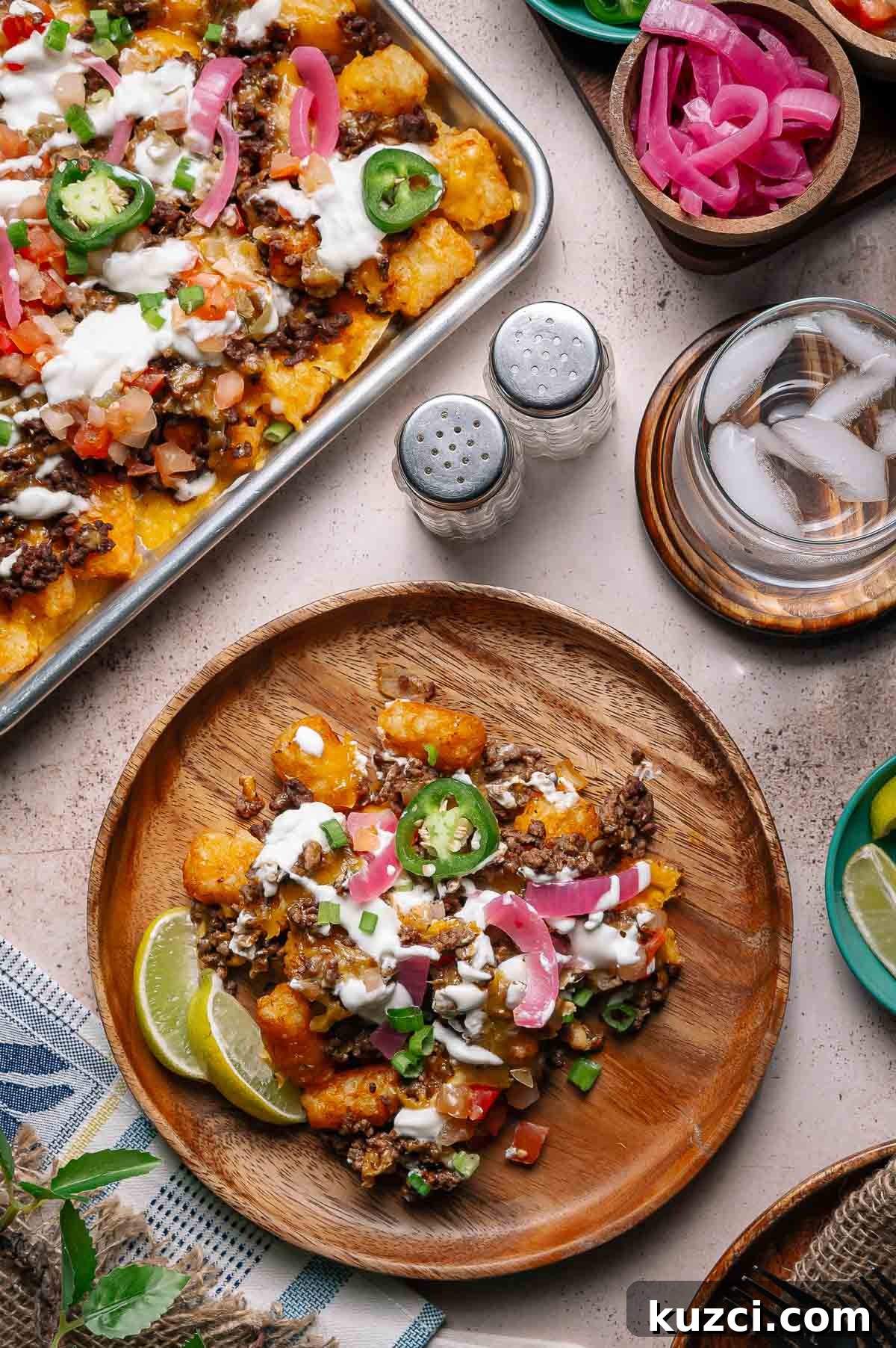 A wooden plate with loaded tater tots topped with ground beef, cheese, jalapeños, pickled onions, sour cream, and lime wedges; a tray of the same dish and condiments are nearby.