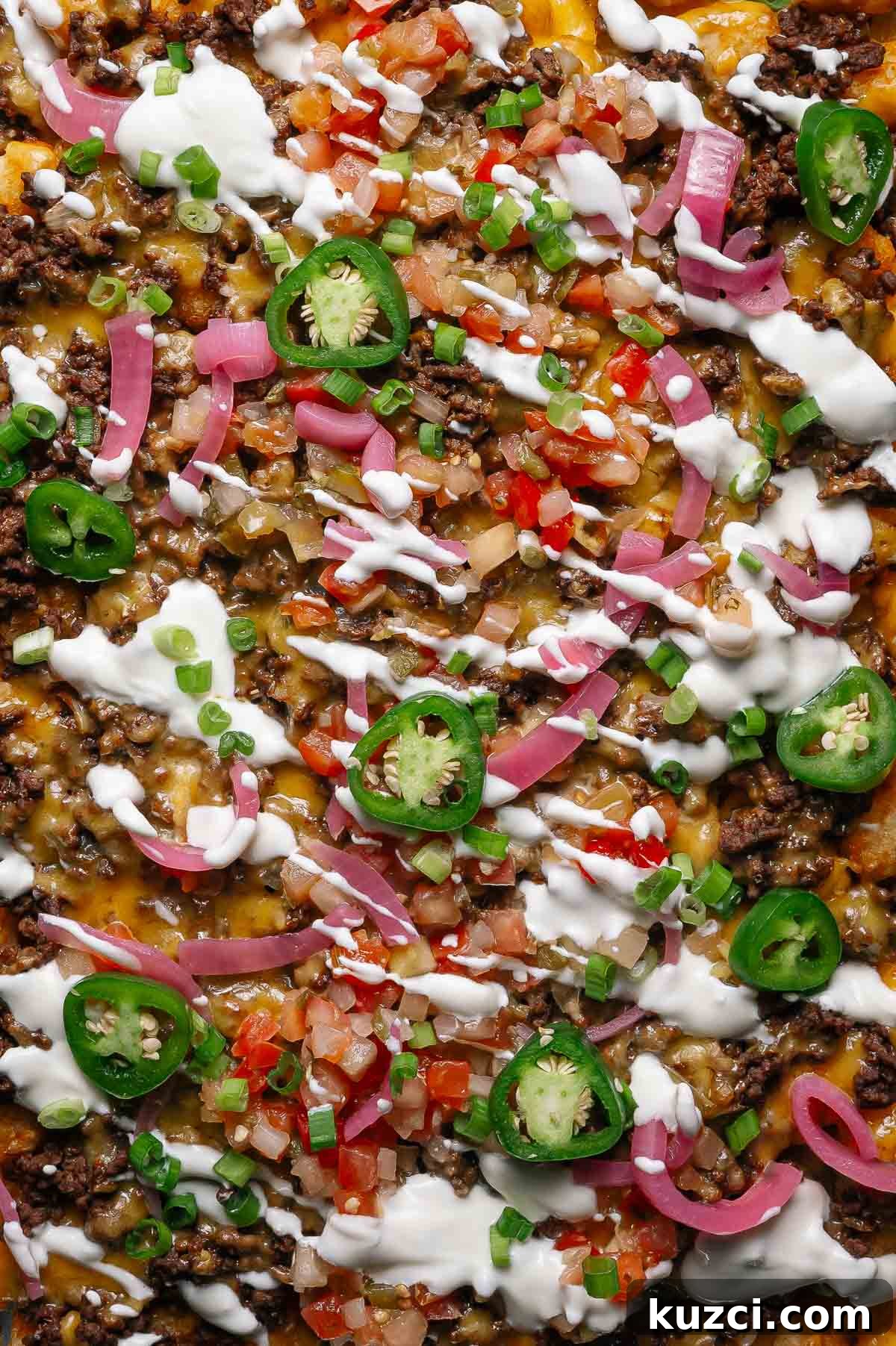 Close-up of totchos topped with ground beef, melted cheese, sliced jalapeños, diced tomatoes, pickled onions, green onions, and drizzled with sour cream.