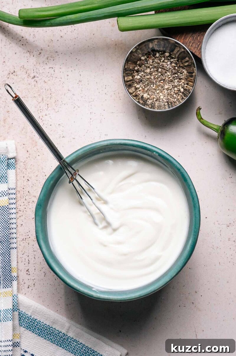 A bowl of lime crema white sauce with a whisk inside, next to a towel, chopped green onions, a jalapeño, ground pepper, and a bowl of salt on a light surface.
