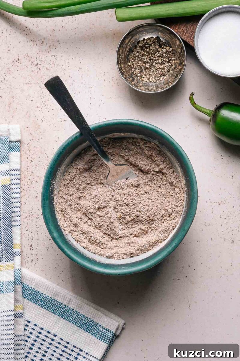 A bowl filled with a dry taco seasoning mixture and a fork, surrounded by a jalapeño, a bowl of pepper, green onions, and a kitchen towel on a countertop.
