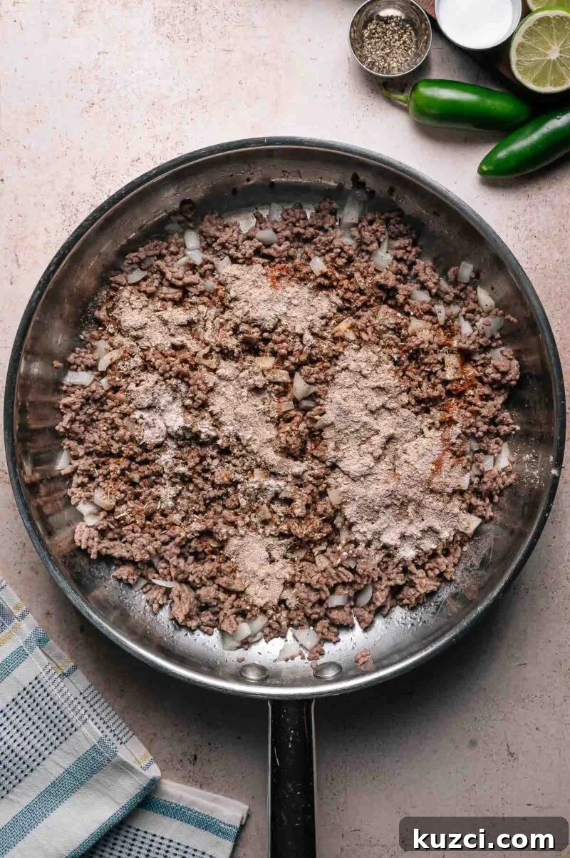 A skillet with cooked ground beef and chopped onions, topped with dry seasoning mix. Nearby are a towel, lime, jalapeños, salt, and pepper.
