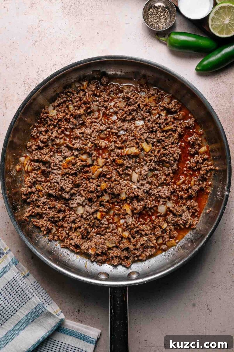 A skillet filled with cooked ground beef mixed with diced onions and spices, placed on a light countertop next to lime, jalapeño, and small bowls of seasoning.
