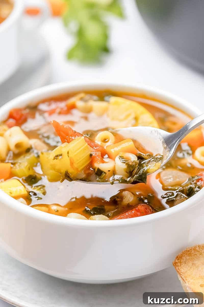 A spoonful of vegetable soup with pasta, celery, carrots, and leafy greens in a white bowl.