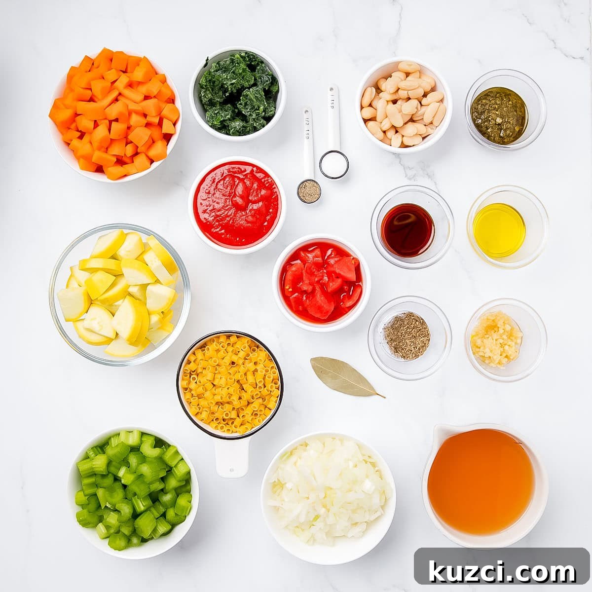 Assorted ingredients for a recipe: diced carrots, kale, white beans, pesto, tomatoes, olive oil, lemon wedges, macaroni, bay leaf, diced celery, chopped onion, broth, minced garlic.