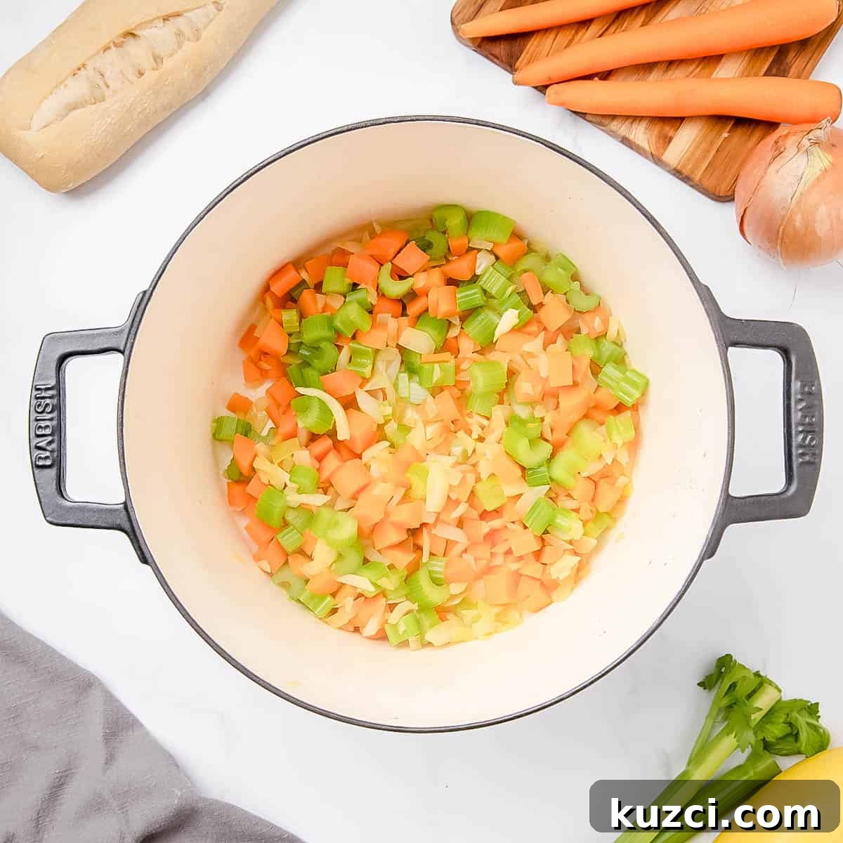 Chopped carrots, celery, and onions are in a pot. Surrounding ingredients include carrots, celery, onion, bread, and a yellow vegetable on a white surface.