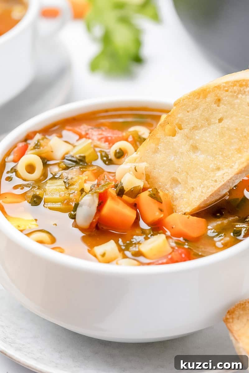 Soup with pasta, carrots, onions, leafy greens, and beans in a white bowl. A piece of baguette is dipped in the center.