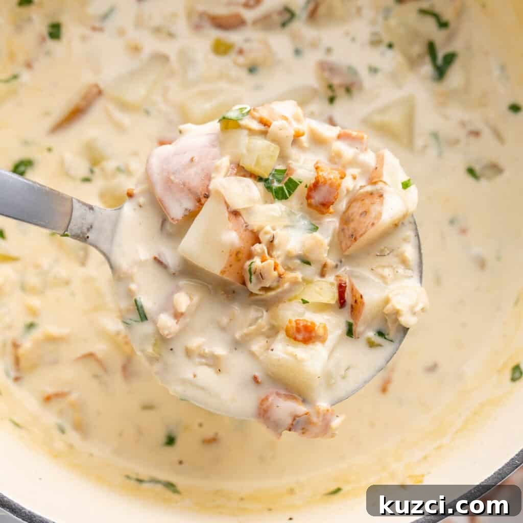 Close-up of a ladle full of creamy clam chowder with diced potatoes, bacon bits, and herbs, over a pot of the soup.