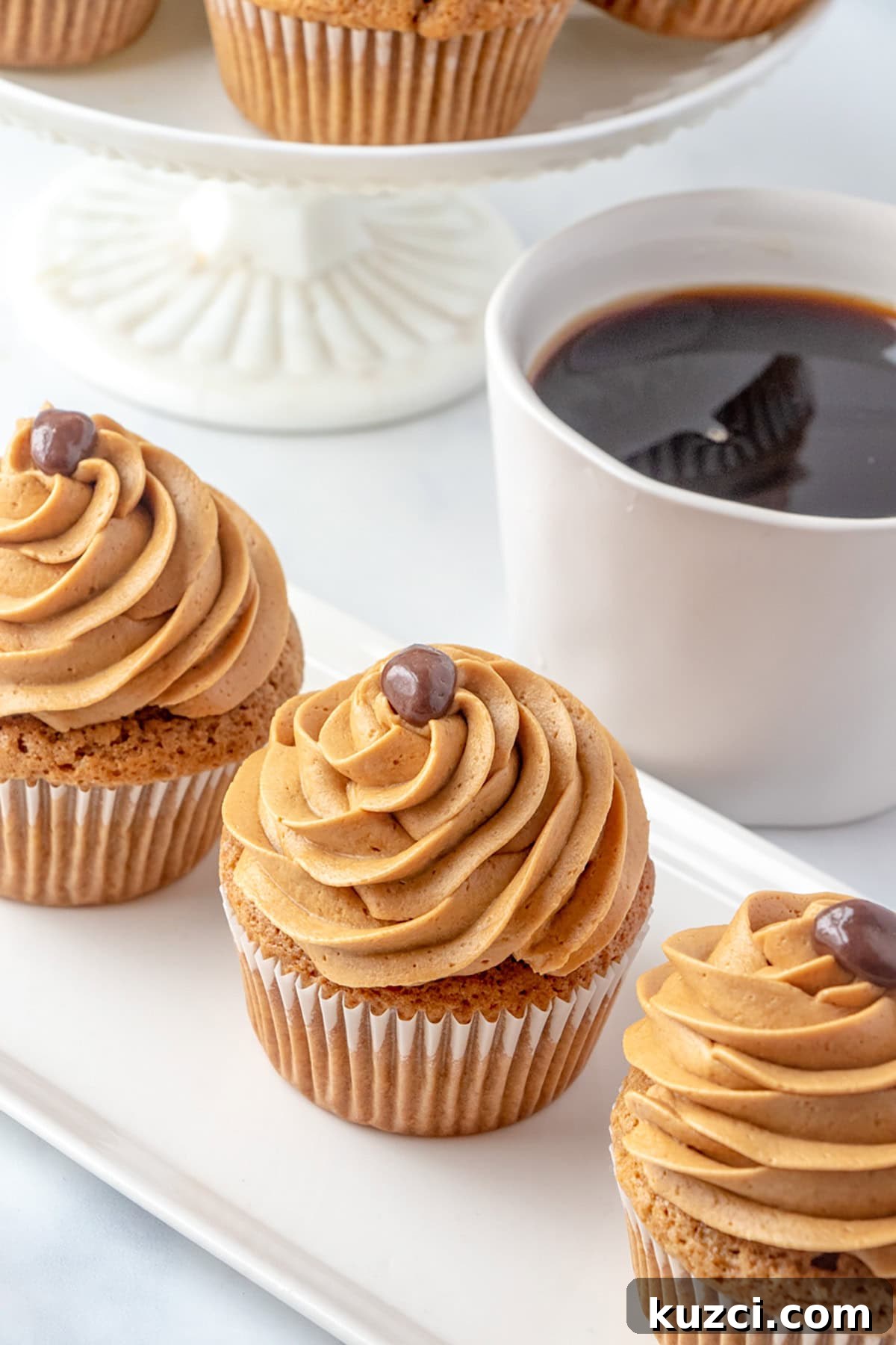 Coffee Cupcake Recipe with Coffee Buttercream Frosting 7 Three Coffee Cupcakes with brown swirled frosting and a small chocolate decoration are arranged on a rectangular plate next to a white mug of black coffee.